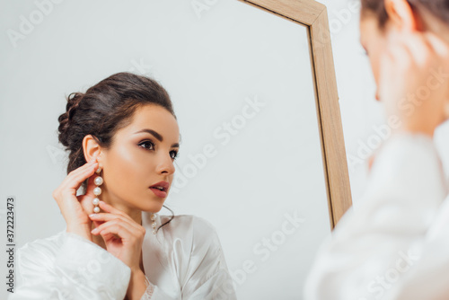 Fotografija Selective focus of bride in silk robe putting on pearl earring near mirror