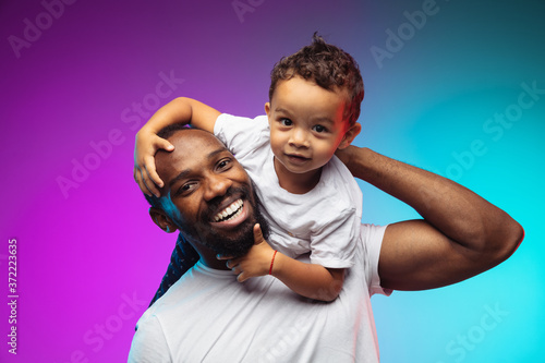 African-american father and son portrait on gradient studio background in neon. Beautiful male models in casual style, white shirt. Concept of human emotions, facial expression, sales, ad, family. photo