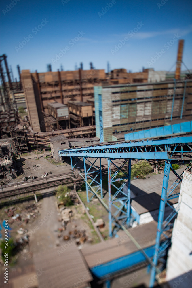 Old metallurgy factory buildings with blue belt conveyor and factory chimneys on blue sky. Birds-eye view and tilt-shift lens.