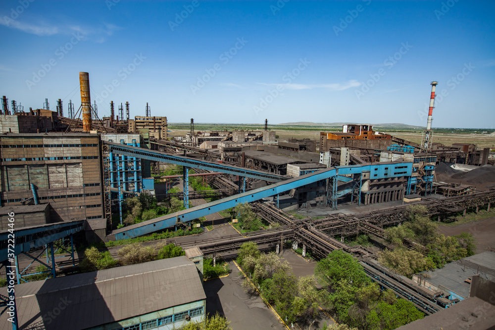Abandoned metallurgy plant (factory) buildings with blue conveyor and rusted chimneys on the blue sky. Panorama aerial view. Taraz city, Kazakhstan.