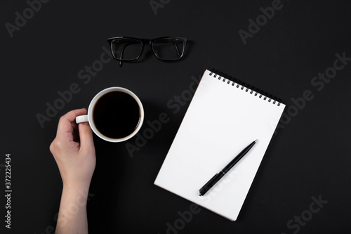  hand holds a cup of coffee on a working black table next to notepad, pen. Top view with copy space for text input.