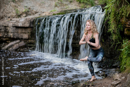 Blonde woman in sportswear practicing yoga at the forest riverside near the waterfall. Fitness girl doing a balance exercise  standing on one leg with hands clasped with in namaste mudra sign