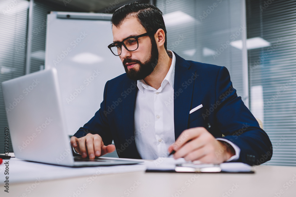 Concentrated male entrepreneur in formal wear watching online database to check accounting reports on modern netbook device and making information notes working in office of financial company