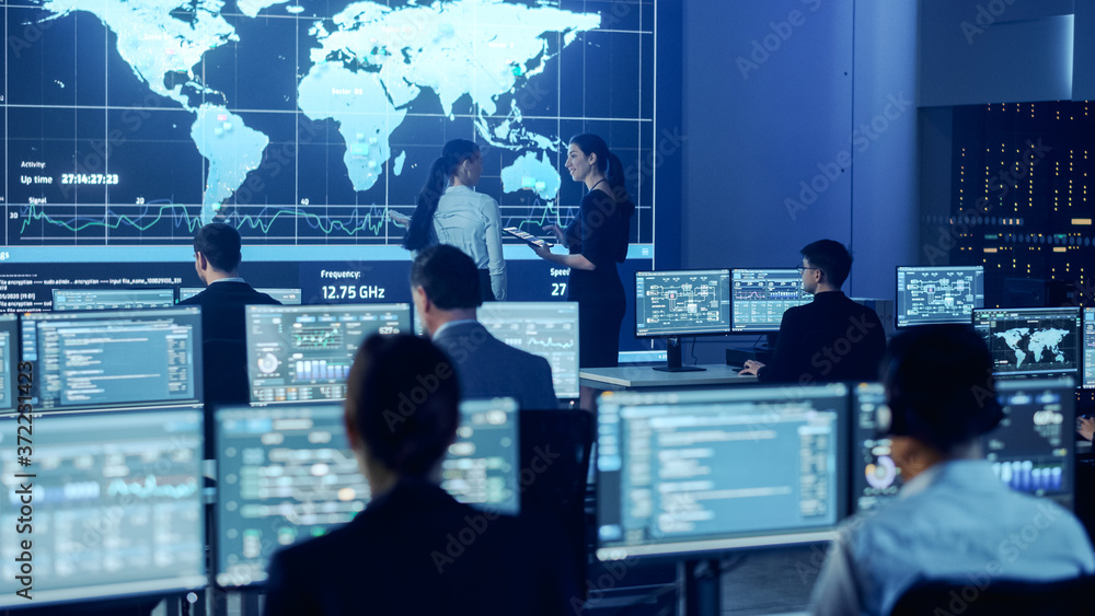Project Manager and Computer Science Engineer Talking while Using Big Screen Display Showing Global Map and Data. Telecommunications Company System Control and Monitoring Room. Project Manager and Computer Science Engineer Talking while Using Big Screen Display Showing Global Map and Data. Telecommunications Company System Control and Monitoring Room.