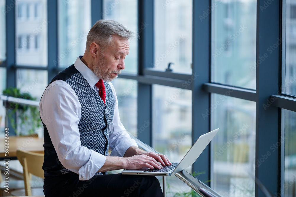 Adult male businessman, teacher, mentor working on a new project. Sits by a large window on the table. He looks at the laptop screen. Adult male businessman, teacher, mentor working on a new project. Sits by a large window on the table. He looks at the laptop screen.