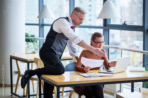 Girl business woman sitting at a wooden table with a laptop and discussing a new project with her mentor boss teacher. The man is fooling around. New business development concept