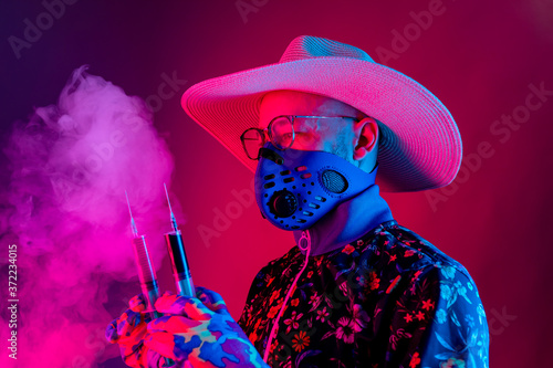 Strange man in medical respirator mask, glasses and hat holding two syrenges with coronavirus covid 19 test on red blue background.with smoke. Evil doctor, inventor of corona virus covid 19 conept photo