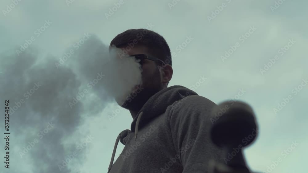 Stylish, fashionable, young guy Caucasian appearance, in sunglasses, smokes hookah from pipe, exhales, releases cloud smoke, looks camera. View from above. On plain white background