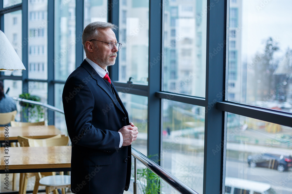 Adult male businessman, teacher, mentor looks out the large window of his office. Work in the office. Development of a new business project Adult male businessman, teacher, mentor looks out the large window of his office. Work in the office. Development of a new business project