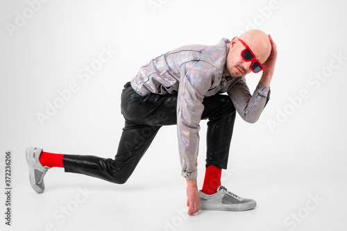 Beautiful caucasian man without hair in grey shirt, black leather trousers, black sunglasses, red socks and grey trainers poses for the camera isolated on white background