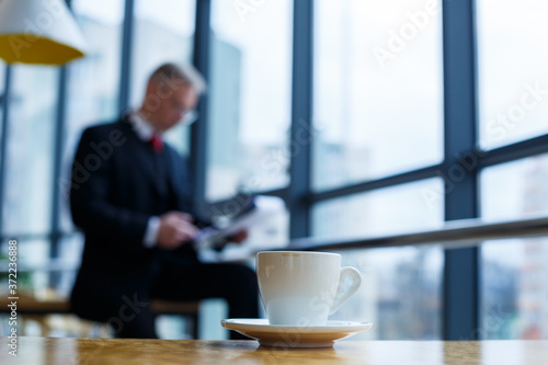 A white cup and saucer with hot aromatic coffee stands on a wooden table against the background of a sitting businessman