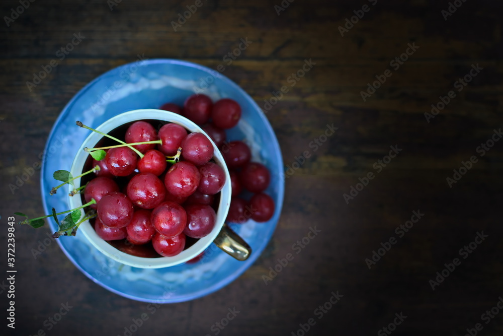Blue cup on saucer with full red healthy ripe sweet cherry berries on old dark wooden background. Top view Blue cup on saucer with full red healthy ripe sweet cherry berries on old dark wooden background. Top view