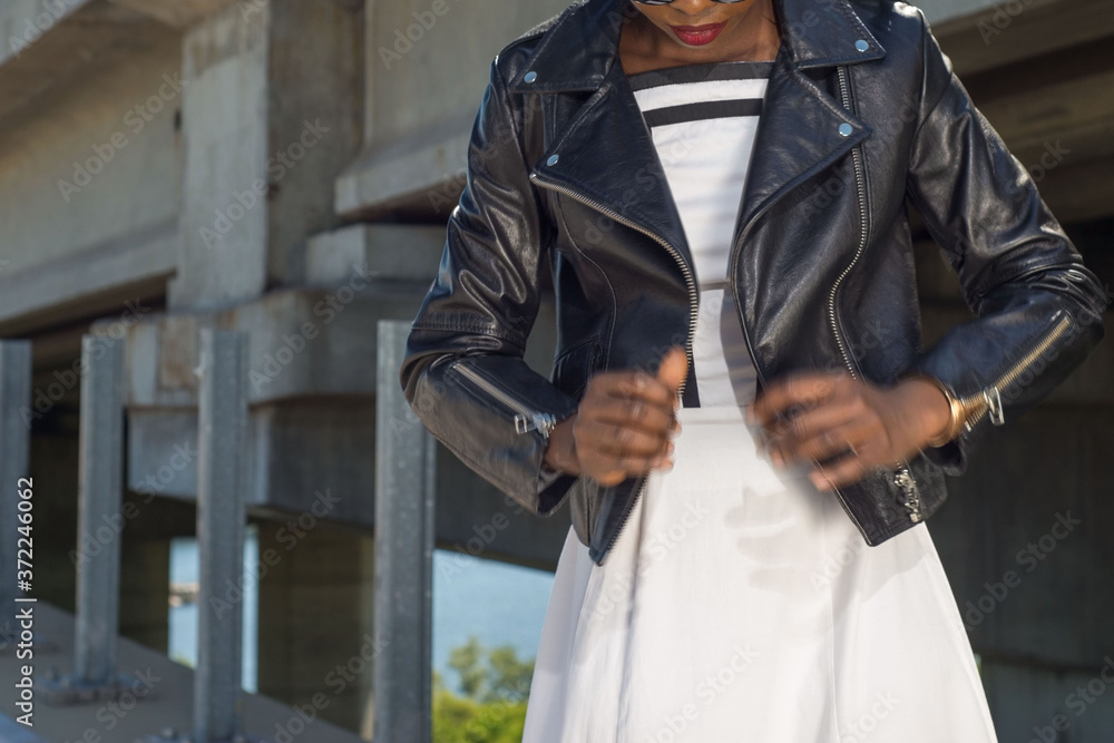 African fashion model, in black leather jacket, white dress, posing. Against the background of street texture