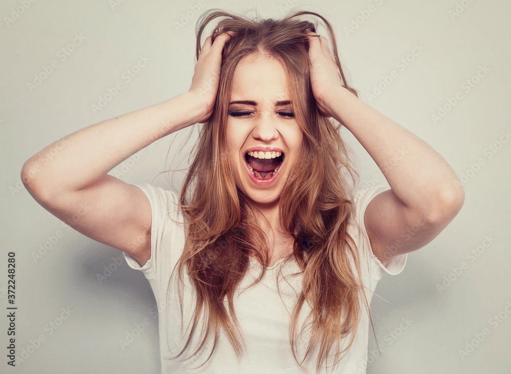 Obraz premium Angry young blong woman strong screaming with wide open mouth on light blue background. Closeup portrait