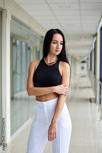 Young brunette woman, wearing white pants and black top, standing in light passageway with huge windows, posing. Businesswoman on a break. Female portrait photography.