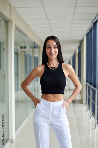 Free Download Young brunette woman, wearing white pants and black top, standing in light passageway with huge windows, posing. Businesswoman on a break. Female portrait photography. - GFXLoad