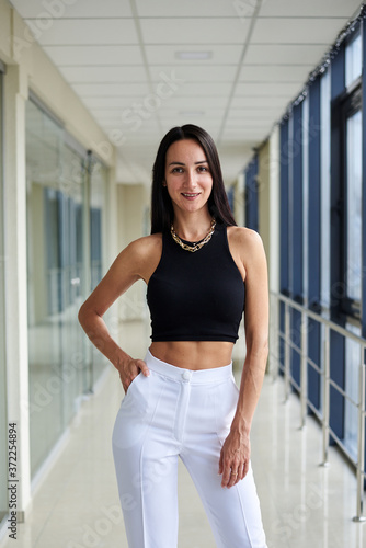 Young brunette woman, wearing white pants and black top, standing in light passageway with huge windows, posing. Businesswoman on a break. Female portrait photography. Free Download Young brunette woman, wearing white pants and black top, standing in light passageway with huge windows, posing. Businesswoman on a break. Female portrait photography. - GFXLoad