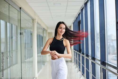 Young brunette woman, wearing white pants and black top, standing in light passageway with huge windows, posing, waving her red hair. Businesswoman on a break. Female portrait photography. Free Download Young brunette woman, wearing white pants and black top, standing in light passageway with huge windows, posing, waving her red hair. Businesswoman on a break. Female portrait photography. - GFXLoad