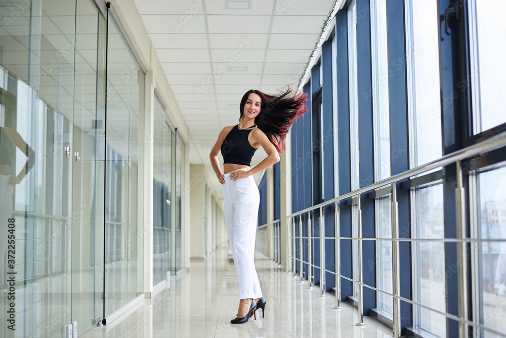 Young brunette woman, wearing white pants and black top, standing in light passageway with huge windows, posing, waving her red hair. Businesswoman on a break. Female portrait photography.