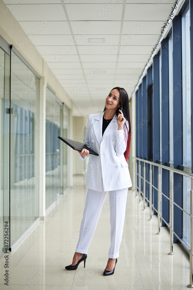 Young brunette woman, wearing white suit, holding black folder with documents,standing in white office building with huge windows.Businesswoman, getting ready for corporate meeting.Business relations