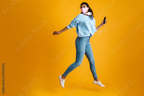 Image of woman in face mask using cellphone and headphones while jumping