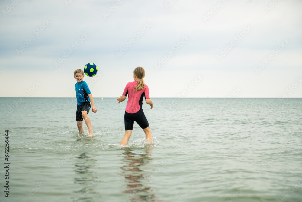 Two happy siblings children playing and jumping with ball in water in neoprene suits in sea Two happy siblings children playing and jumping with ball in water in neoprene suits in sea