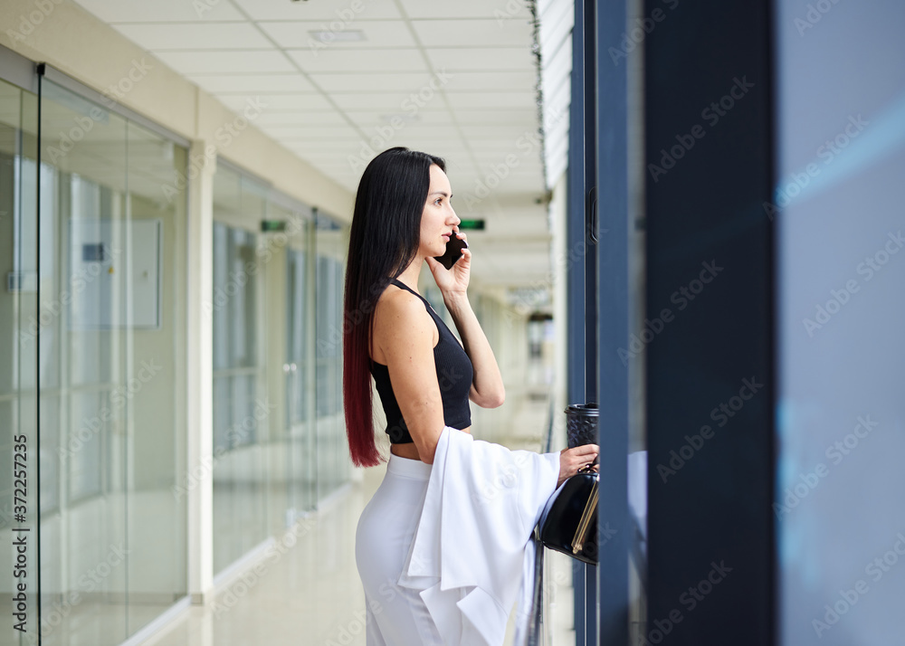 Young brunette woman, wearing white pants and black top, talking on phone, holding coffee cup and purse, standing by the window in light office building. Office worker on break.