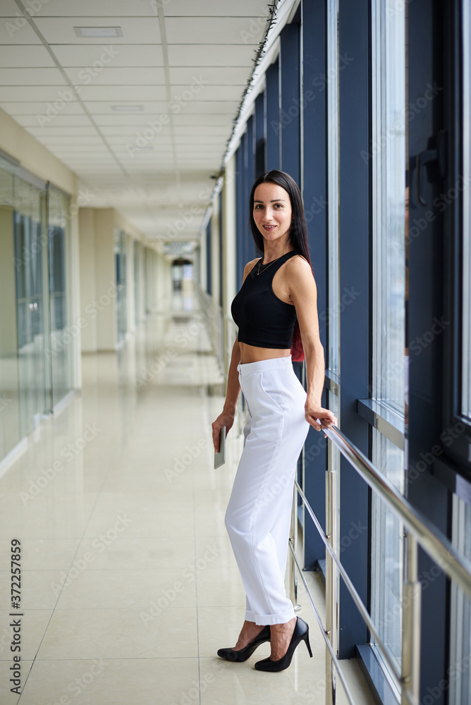 Young brunette woman, wearing white pants and black top, holding silver tablet, standing in light office building. Businesswoman, getting ready for corporate meeting. Office worker on break.