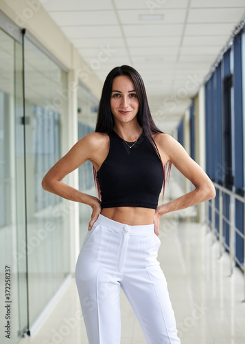 Young brunette woman, wearing white pants and black top, standing in light passageway with huge windows, posing with arms on her waist. Businesswoman on a break. Female portrait photography.