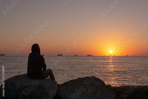 The silhouette of a hooded man sitting on the rocks on the beach at sunset © Денис Кузнецов