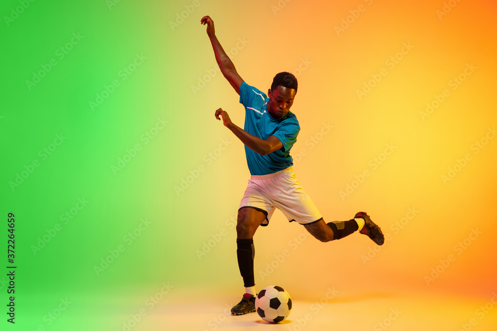 Leader. African-american male soccer, football player training in action isolated on gradient studio background in neon light. Concept of motion, action, ahievements, healthy lifestyle. Youth culture. Leader. African-american male soccer, football player training in action isolated on gradient studio background in neon light. Concept of motion, action, ahievements, healthy lifestyle. Youth culture.