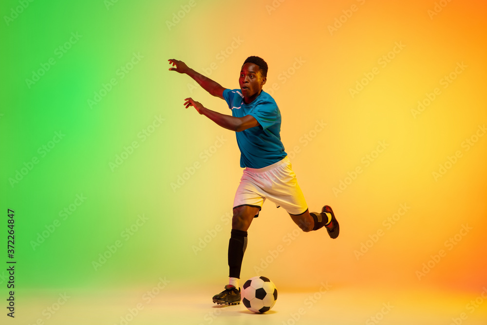 Leader. African-american male soccer, football player training in action isolated on gradient studio background in neon light. Concept of motion, action, ahievements, healthy lifestyle. Youth culture.