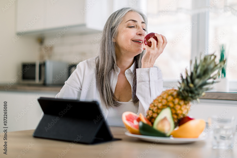 Beautiful elderly female pensioner with long gray hair sitting at the kitchen table at home and enjoying her lunch time with fresh apple and fruits. Tablet computer on the table Beautiful elderly female pensioner with long gray hair sitting at the kitchen table at home and enjoying her lunch time with fresh apple and fruits. Tablet computer on the table