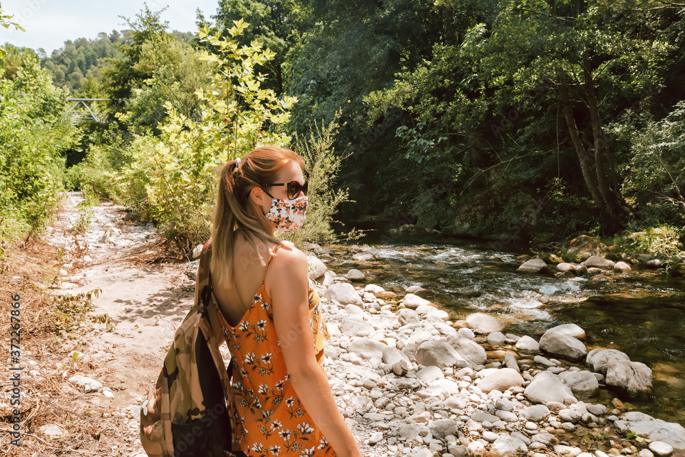 Woman in yellow sundress enjoying a walk near river in face mask due to new normal rules and restrictions. Solo traveler outdoors with backpack, view from behind