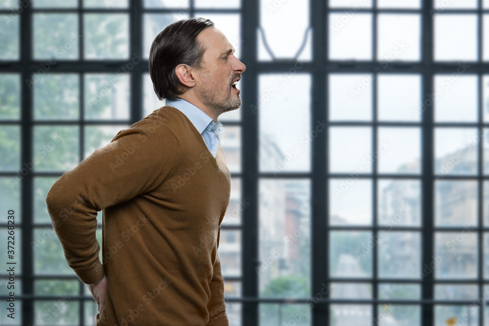 Mature caucasian man suffering from backache. Side view of stressed male employee suffering from back pain. Office windows background. Mature caucasian man suffering from backache. Side view of stressed male employee suffering from back pain. Office windows background.