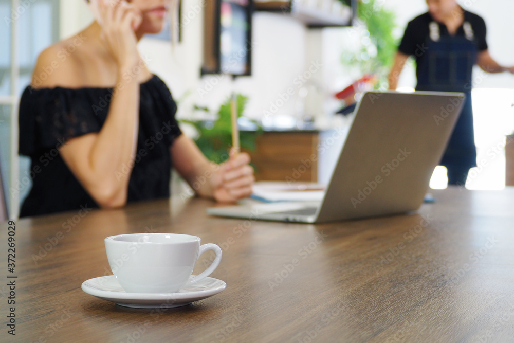 Selective focus on coffee cup and saucer with blur woman working on her computer and talking on the phone at coffee shop. Working lifestyle at cafe with drink concept Selective focus on coffee cup and saucer with blur woman working on her computer and talking on the phone at coffee shop. Working lifestyle at cafe with drink concept