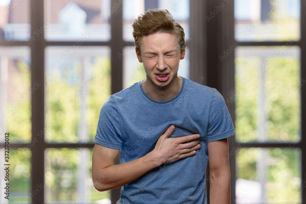 Young caucasian boy suffering from sudden heart attack. Stressed teen guy having chest pain on windows background. Health problem concept. Young caucasian boy suffering from sudden heart attack. Stressed teen guy having chest pain on windows background. Health problem concept.