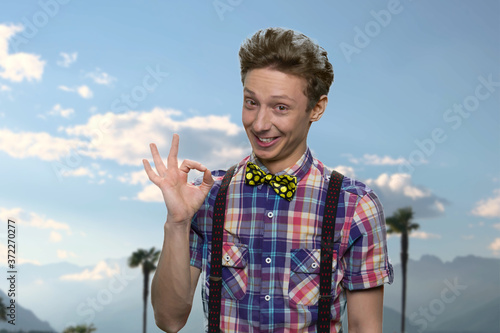 Cute happy teenage boy showing ok sign. Funny teen guy giving OKAY gesture on blue sky background. Summer vacation at tropical resort.
