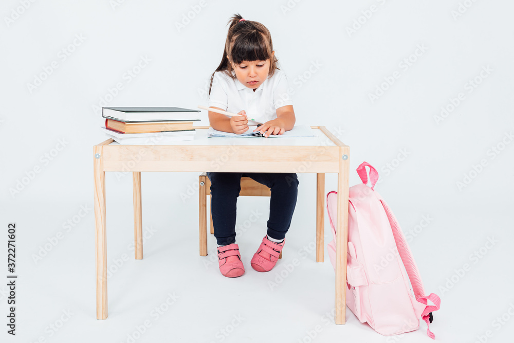 Fototapeta premium Brunette girl at school, smiling sitting on a chair next to a table with books painting, on white background. School concept