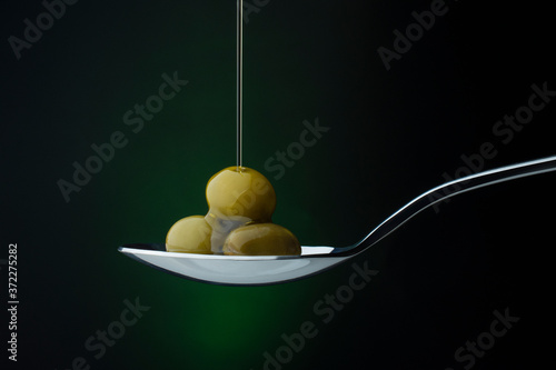 Photo green olives on a spoon on a dark background, stream of olive oil