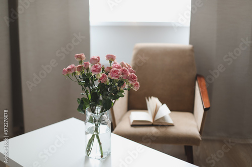 Modern room interior with armchair, book and fresh roses on table. Interior of beautiful living room decorated with flowers