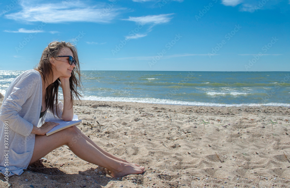 Happy woman reads on the beach. European brunette woman in sunglasses reading a book by the sea and looking at the horizon