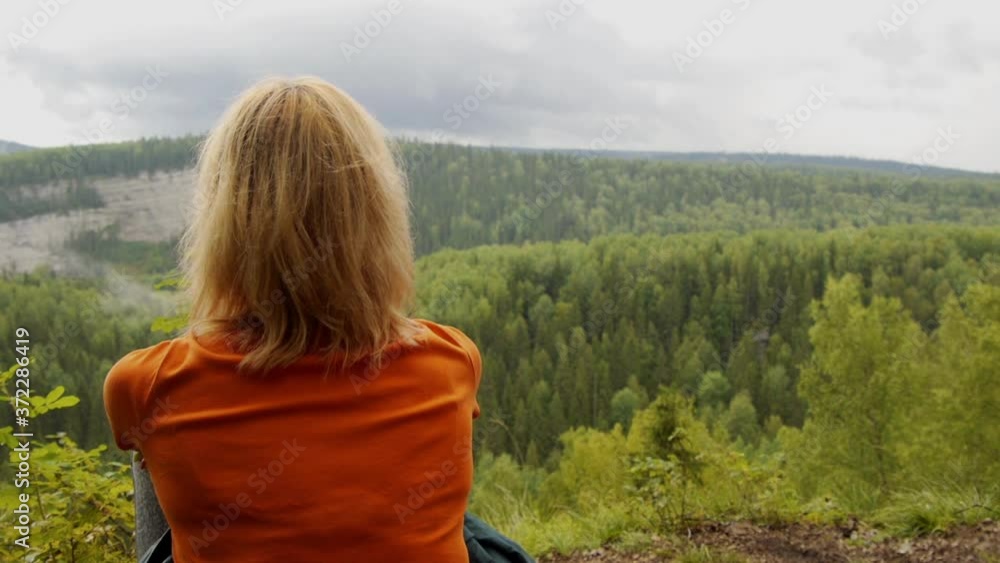 A blonde girl sits on the top of a high mountain and looks down at the panorama of the forest and rocky mountains below A blonde girl sits on the top of a high mountain and looks down at the panorama of the forest and rocky mountains below