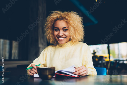 Portrait of cheerful female manager creating checklist in notepad enjoying rest in cafe looking at camera, positive young student with curly blonde hair making daily schedule during coffee break