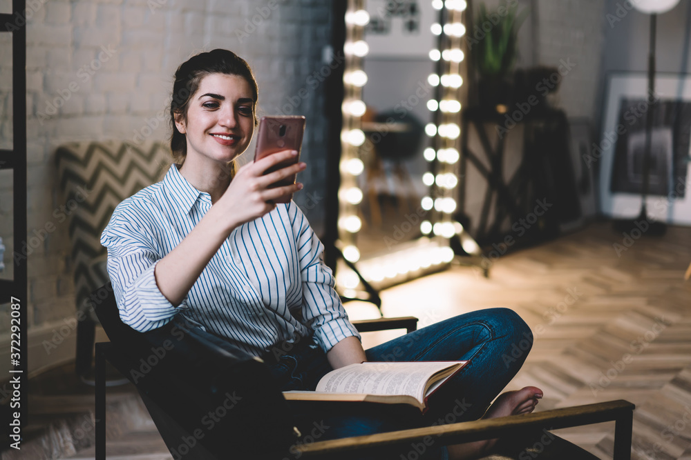 Fototapeta premium Positive young woman resting with book on chair at home stylish interior while reading notification with good news on smartphone.Cheerful female blogger watching funny video in blog enjoying free time
