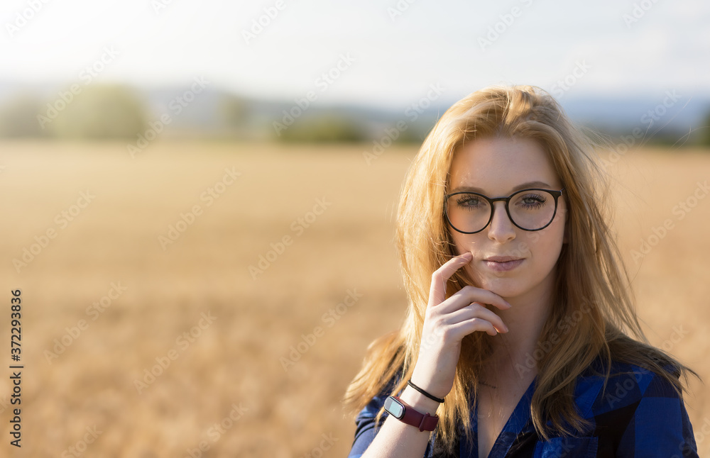 Portrait of young woman with glasses wearing a blue plaid shirt is pensively posing in corn field. Horizontally. 