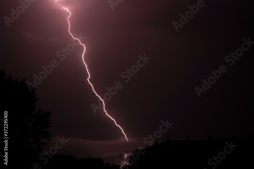 A very branching lightning flashes above the roofs of city houses in a summer thunderstorm. Lightning in the sky over the night city.