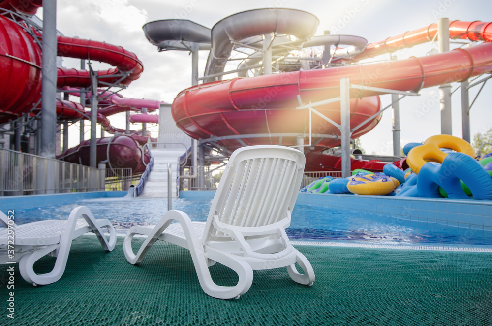 white loose sun loungers near the pool against the background of multi-colored plastic slides in the water park under the sunlight