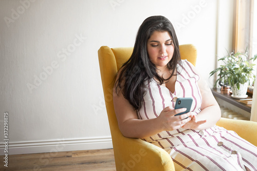 Bild auf Leinwand Pretty girl in plus size dress sitting in armchair with mobile, reading sms from boyfriend