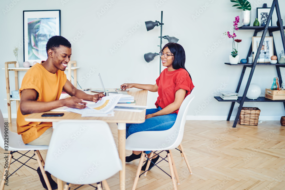 Obraz premium Cheerful african american colleagues sitting at desktop smiling and communicating about startup project, smiling dark skinned marriage planning family budget at table in bright interior living room.
